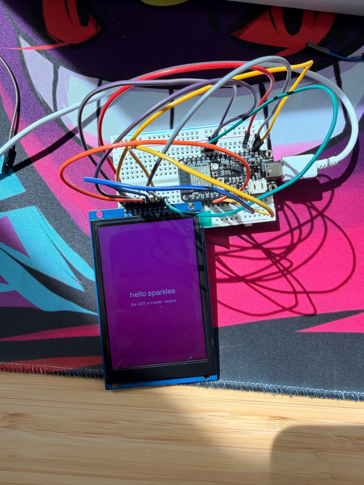 The 3.5-inch display rendering 'hello sparkles β the AO3 e-reader begins' in white text on a pink-purple background, wired to the Feather on the breadboard